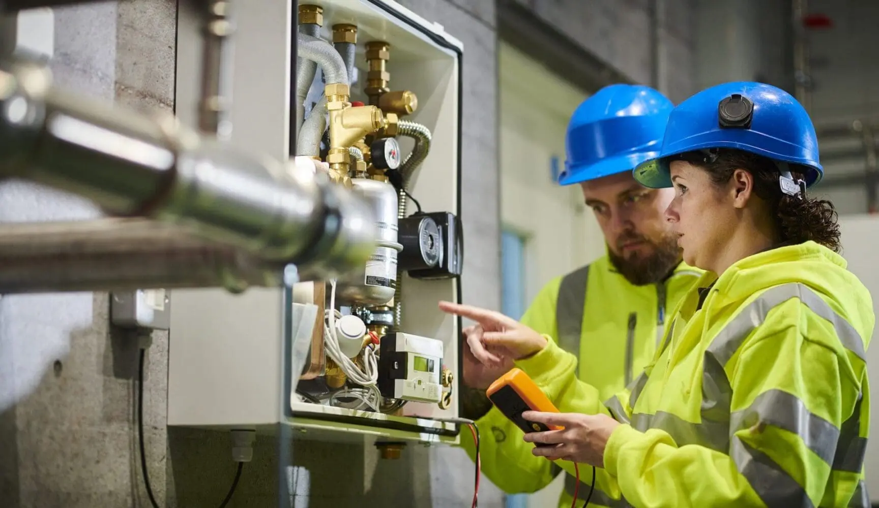 Adherent (formerly Compliance & Risks). Two technicians in blue hard hats and high-visibility jackets inspect an open utility panel filled with pipes, valves, and wiring. One points while the other holds a digital multimeter.