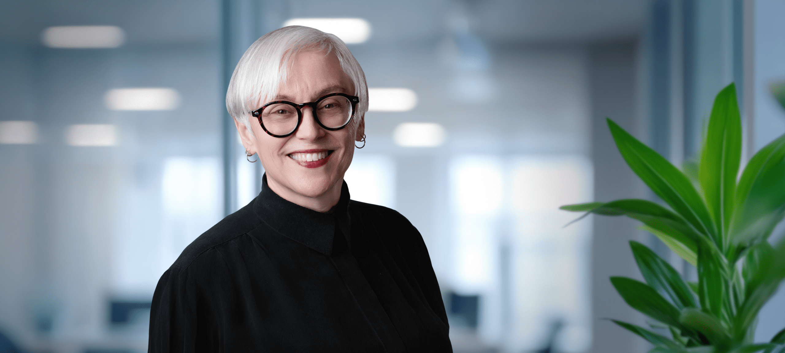 Adherent (formerly Compliance & Risks). Headshot of a smiling woman with short gray hair, round tortoiseshell glasses, and a black shirt, against a blurred office background with a green plant.