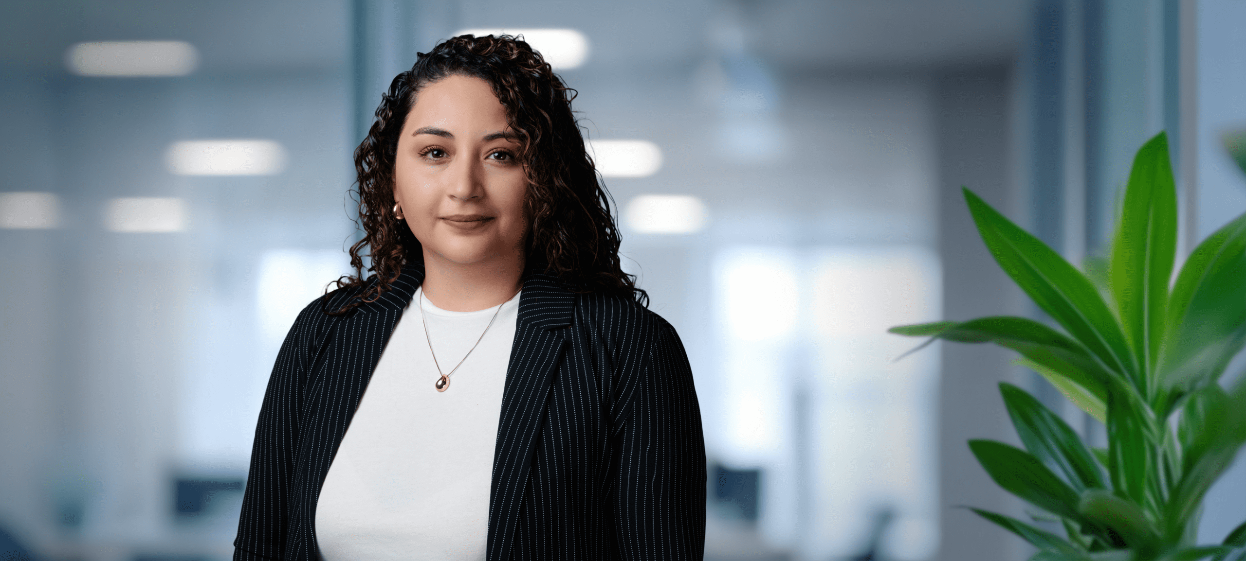 Adherent (formerly Compliance & Risks). A woman with dark curly hair, wearing a pinstripe blazer and a gold necklace, smiles confidently at the camera in a blurred office setting.