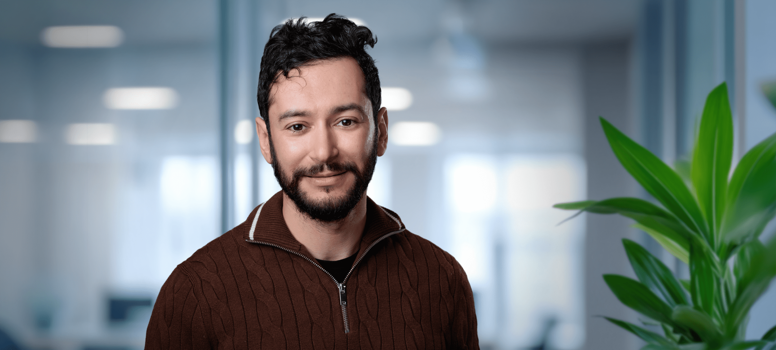 Adherent (formerly Compliance & Risks). A man with dark curly hair and a beard smiles at the camera, wearing a brown cable-knit sweater, with a blurred office background and a green plant on the right.