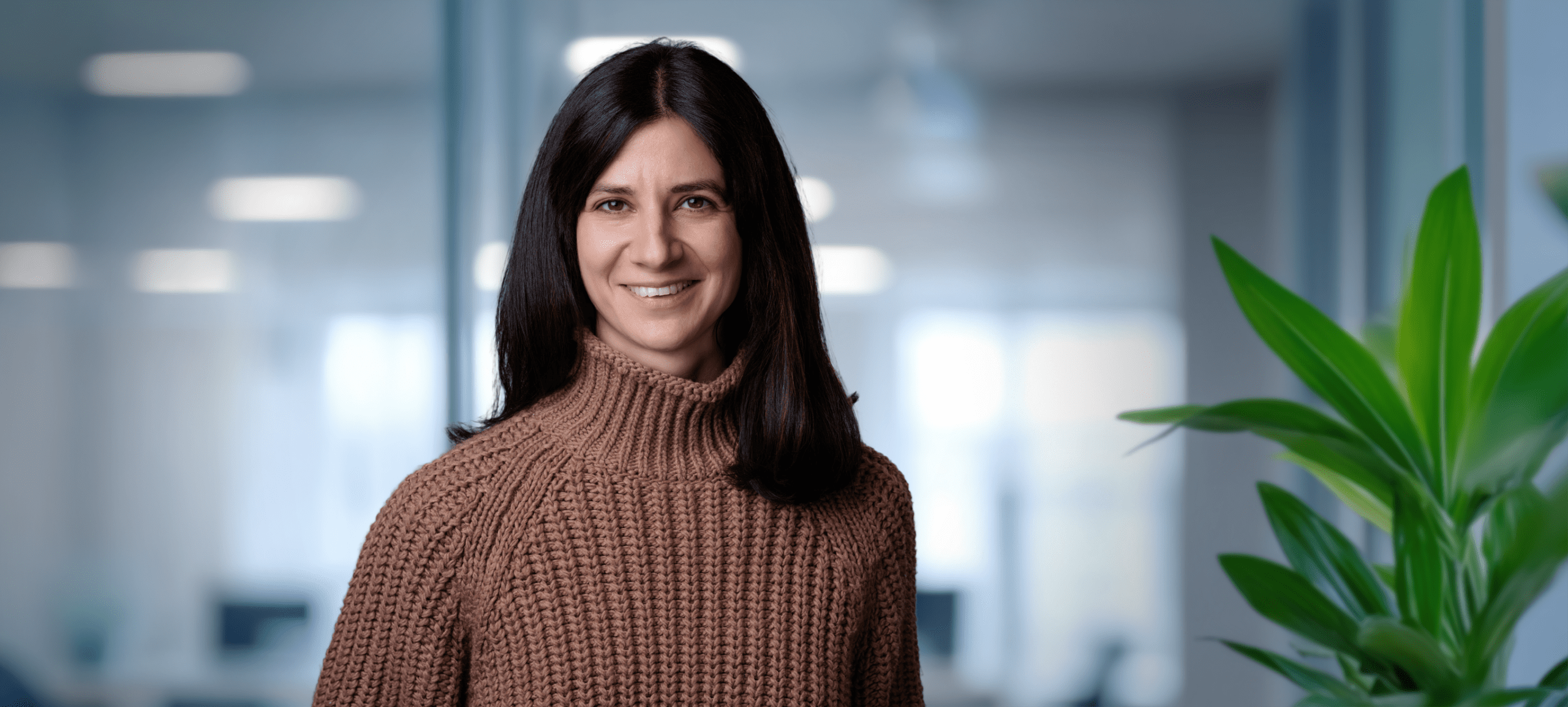 Adherent (formerly Compliance & Risks). A smiling woman with long dark hair in a brown knit sweater, against a blurred office background with a green plant.