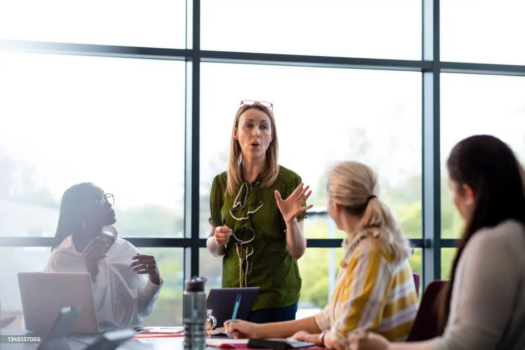 Four women in a bright meeting room. One woman stands, speaking and gesturing, while three others sit at a table, listening attentively. Laptops and notes are on the table.