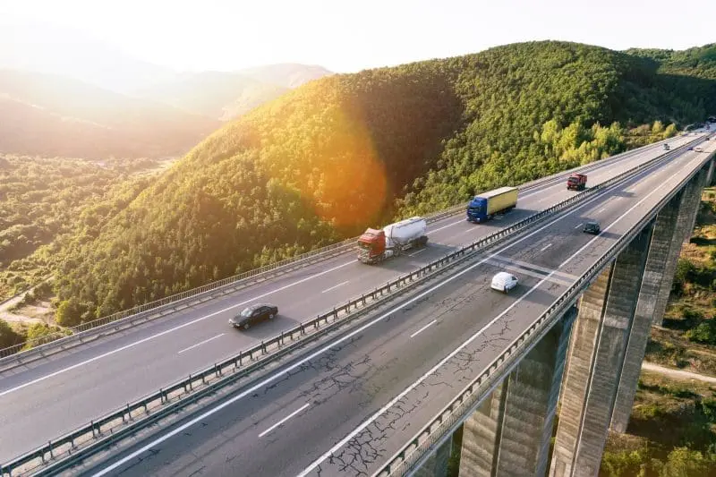 An aerial view of a multi-lane highway bridge spanning a valley between green, forested mountains. Several cars and semi-trucks are driving on the bridge, with a bright sun flare in the upper left.