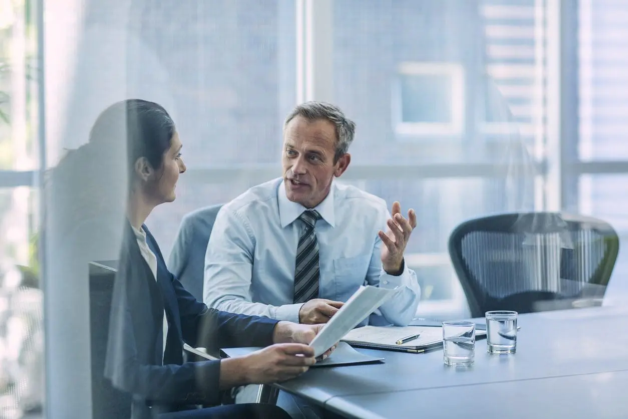 Two business professionals, a man and a woman, discuss documents at a conference table in a bright office with large windows. The man is speaking and gesturing, while the woman listens and holds papers.