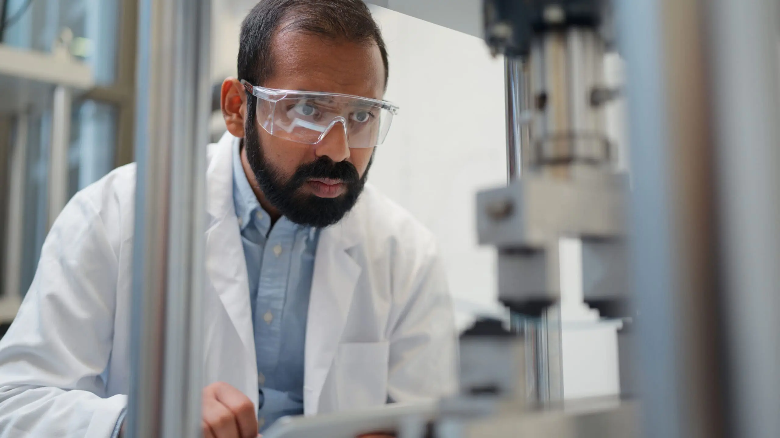 A man in a white lab coat and safety glasses intently looking at laboratory equipment.