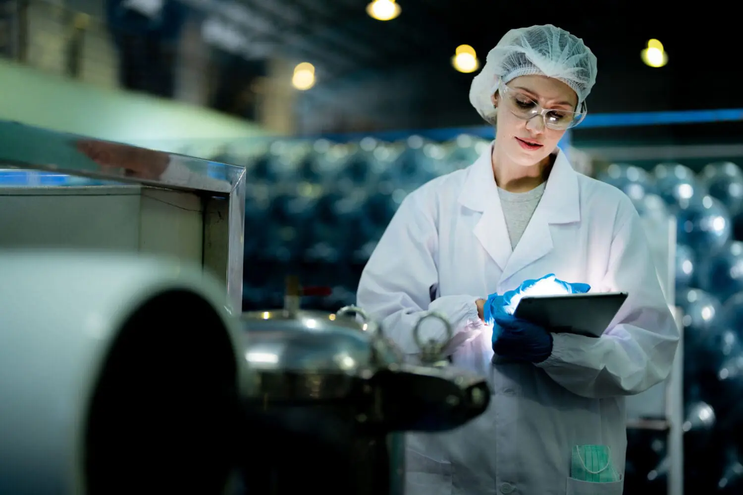 Adherent (formerly Compliance & Risks). A woman in a lab coat, hairnet, and safety glasses looks at a glowing tablet in an industrial setting.