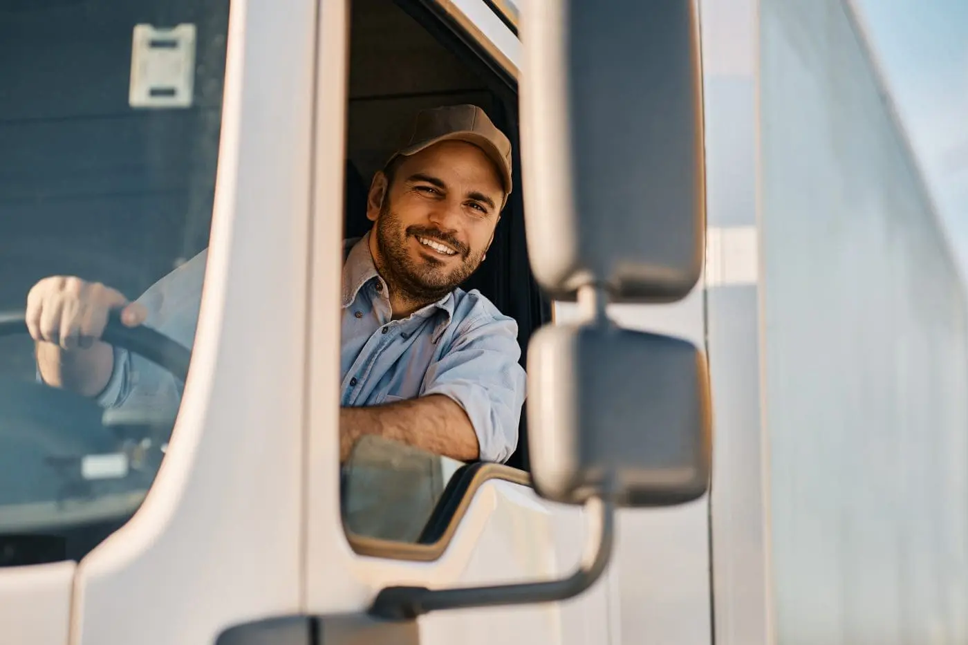 A smiling young male truck driver with a beard and baseball cap leans out of his white truck's window, looking at the viewer.
