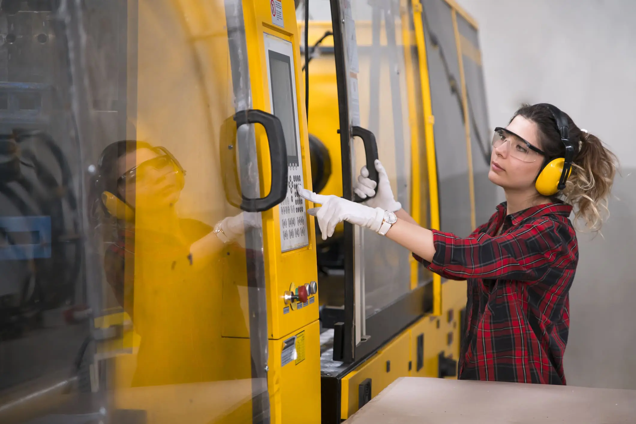 A woman in a red plaid shirt, safety glasses, and yellow ear defenders operates a large yellow industrial machine, pressing buttons on its control panel with gloved hands.