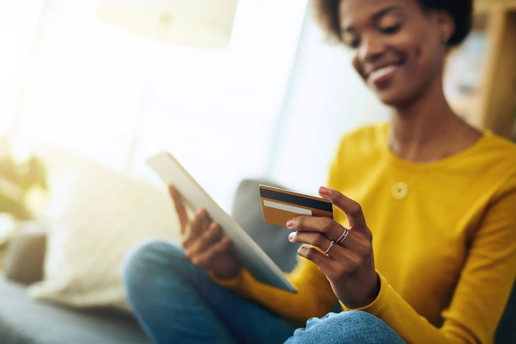 Adherent (formerly Compliance & Risks). A smiling woman in a yellow top and jeans holds a tablet and a gold credit card, appearing to make an online purchase. Bright light illuminates the scene.