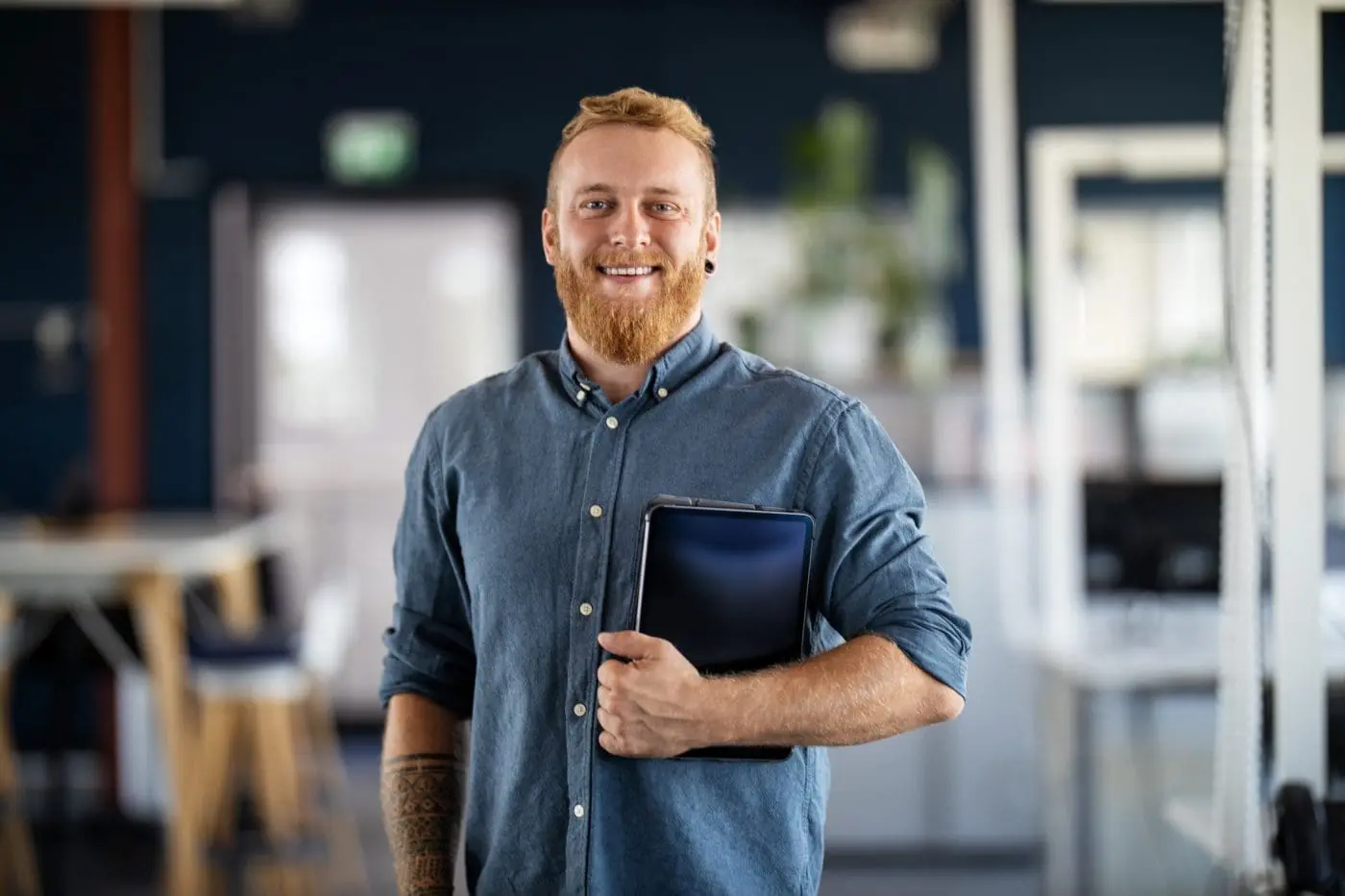 A smiling man with a red beard and a blue shirt holds a tablet in a modern office.