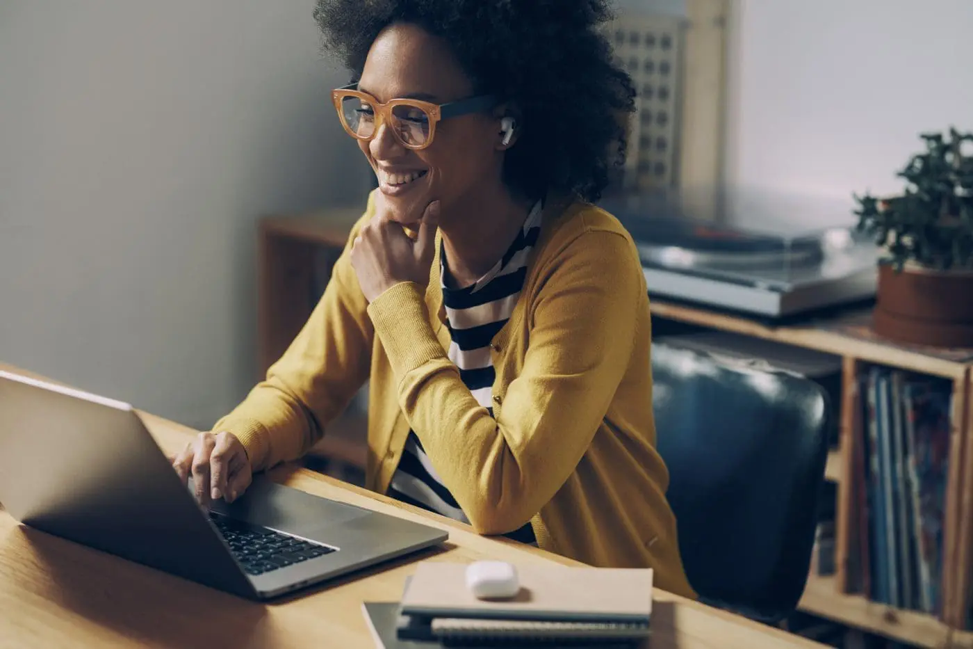 A smiling Black woman with curly hair and orange glasses works on a laptop at a wooden desk, wearing a yellow cardigan.