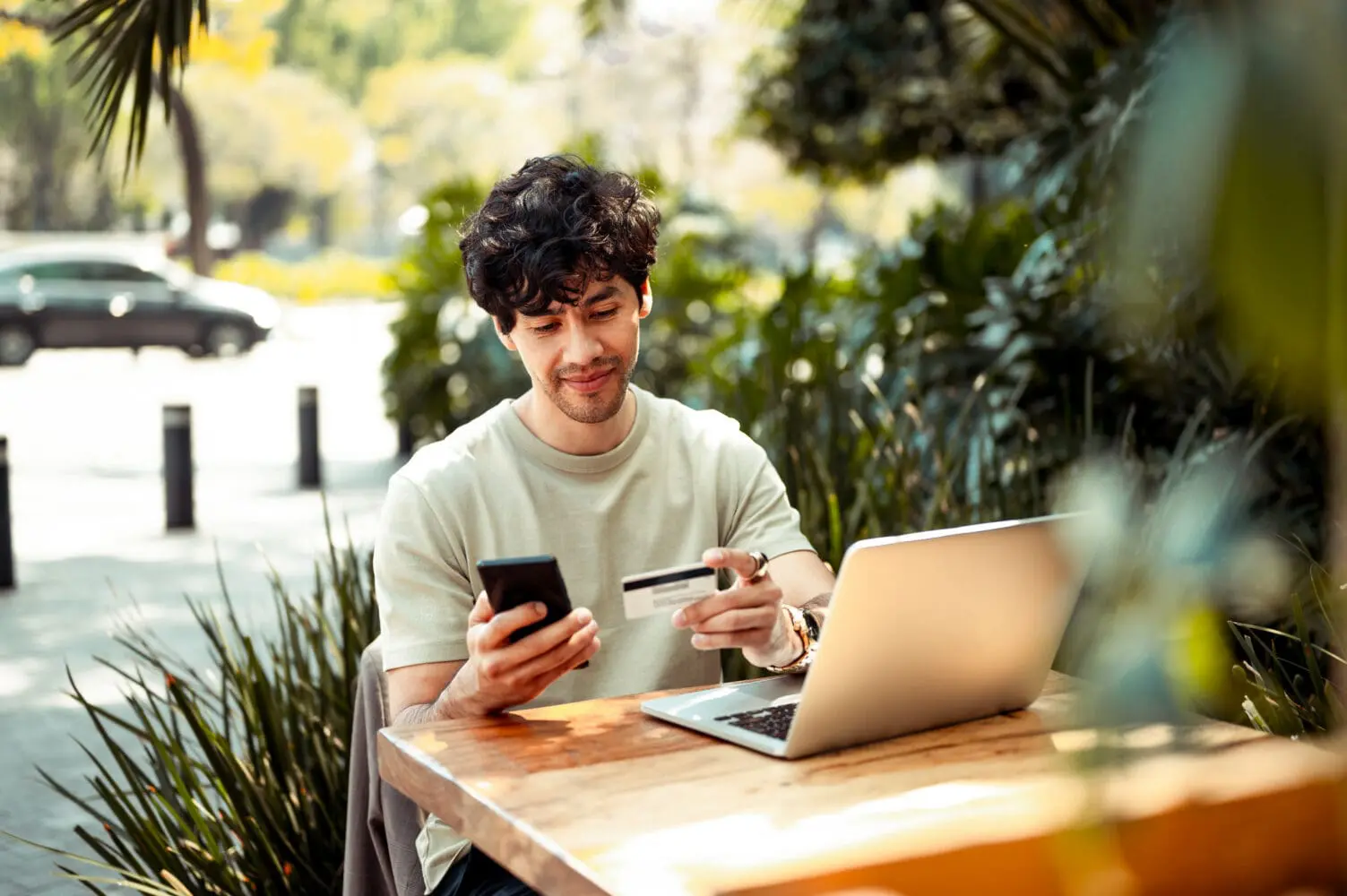 Adherent (formerly Compliance & Risks). A young man with curly hair sits at a wooden table outdoors, holding a smartphone in one hand and a credit card in the other, with a laptop open beside him. He is focused on his devices.