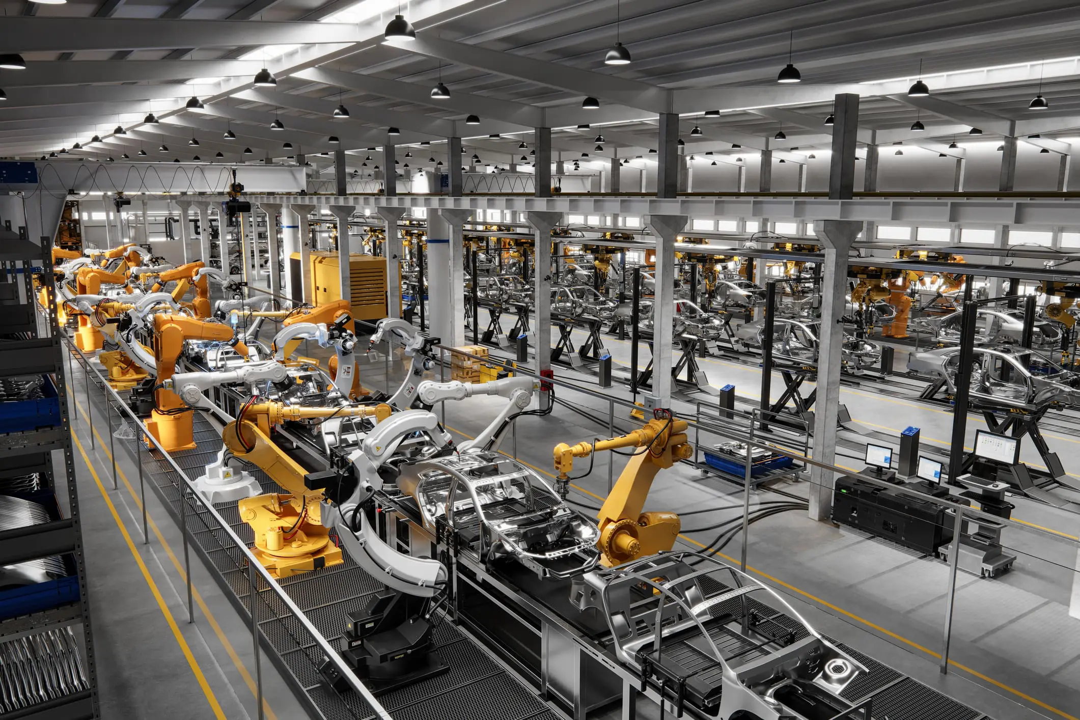 Overhead view of a highly automated car manufacturing plant, featuring numerous yellow and white robotic arms assembling silver car chassis on multiple production lines.