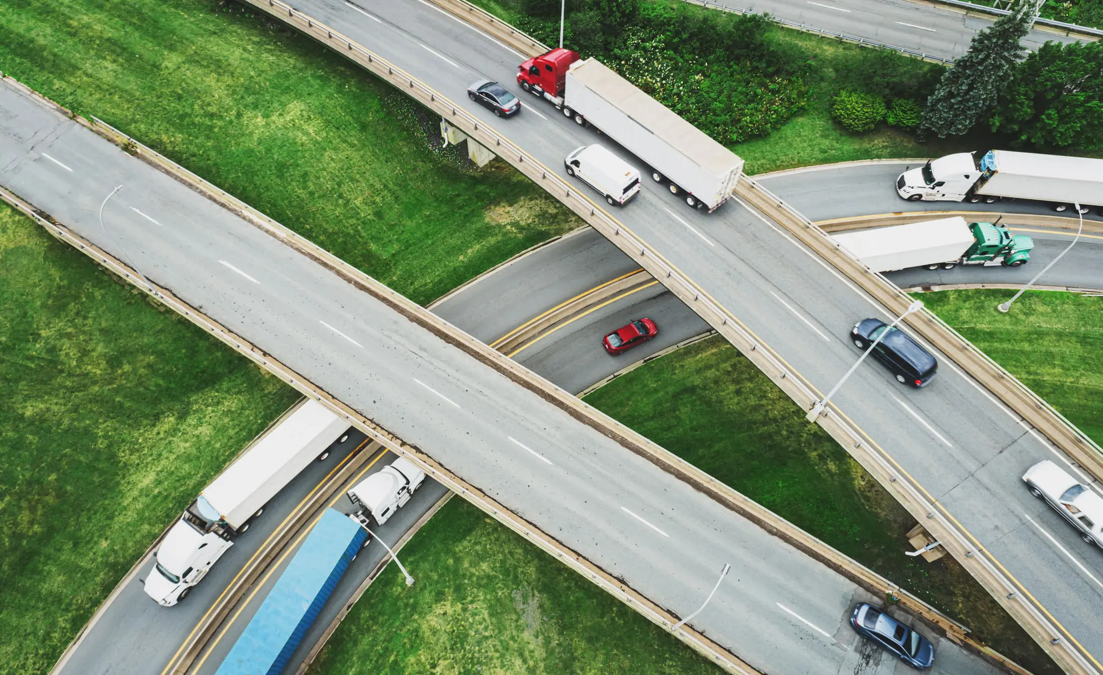 Aerial view of a multi-level highway interchange with cars and semi-trucks on gray roads surrounded by green grass.