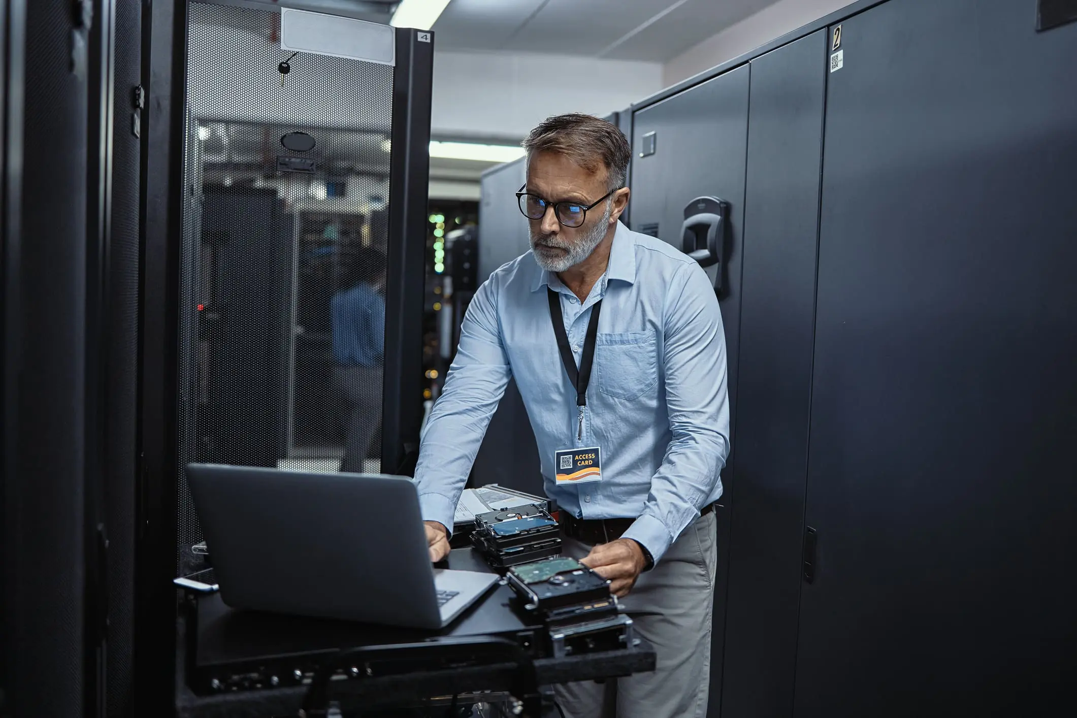 A man with glasses and a beard, wearing a blue shirt, works on a laptop with several hard drives in a data center.