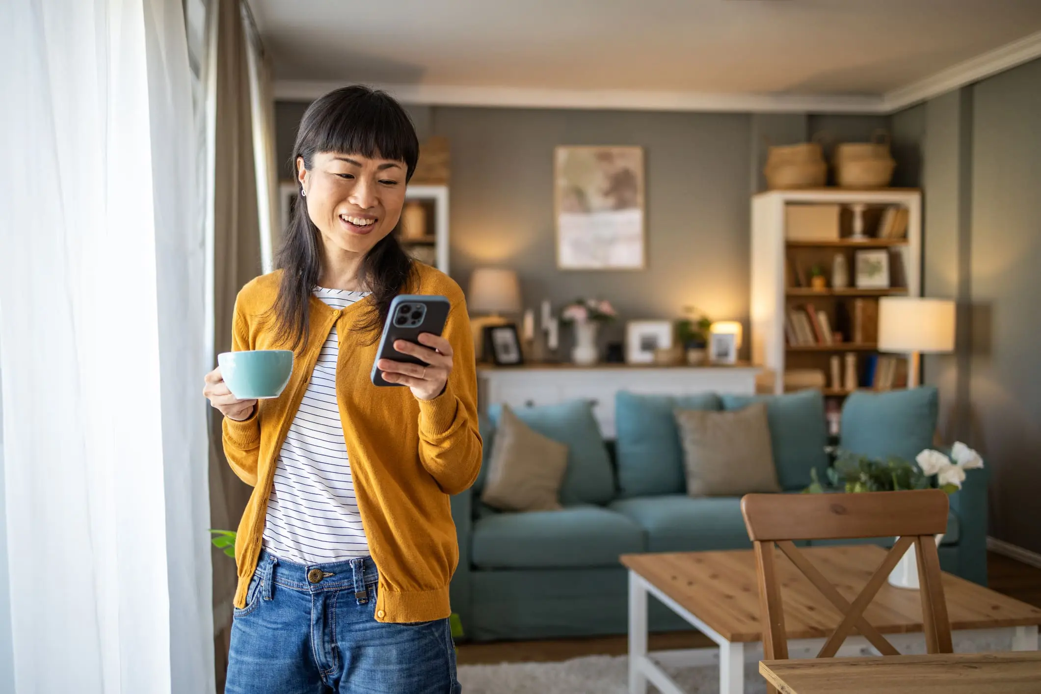 Adherent (formerly Compliance & Risks). A smiling East Asian woman in a yellow cardigan holds a blue mug and looks at her smartphone in a cozy living room.