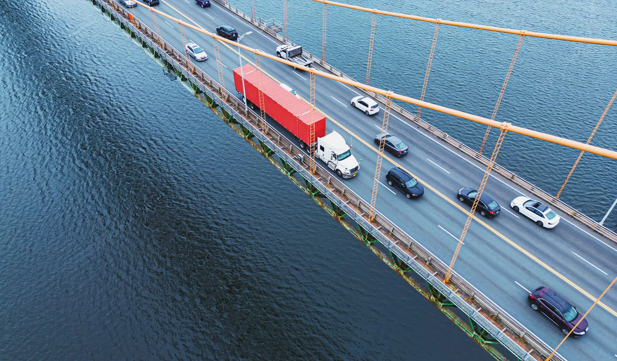 Adherent (formerly Compliance & Risks). High-angle view of a suspension bridge over dark water. Cars and a large red semi-truck are driving on the bridge, supported by orange cables.