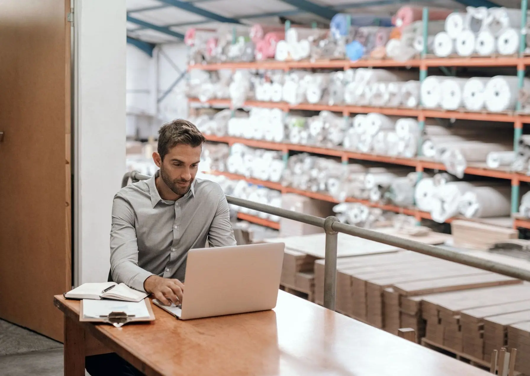 A man in a grey shirt works on a laptop at a wooden desk in a warehouse, with shelves full of rolled materials and stacked boxes visible in the background.