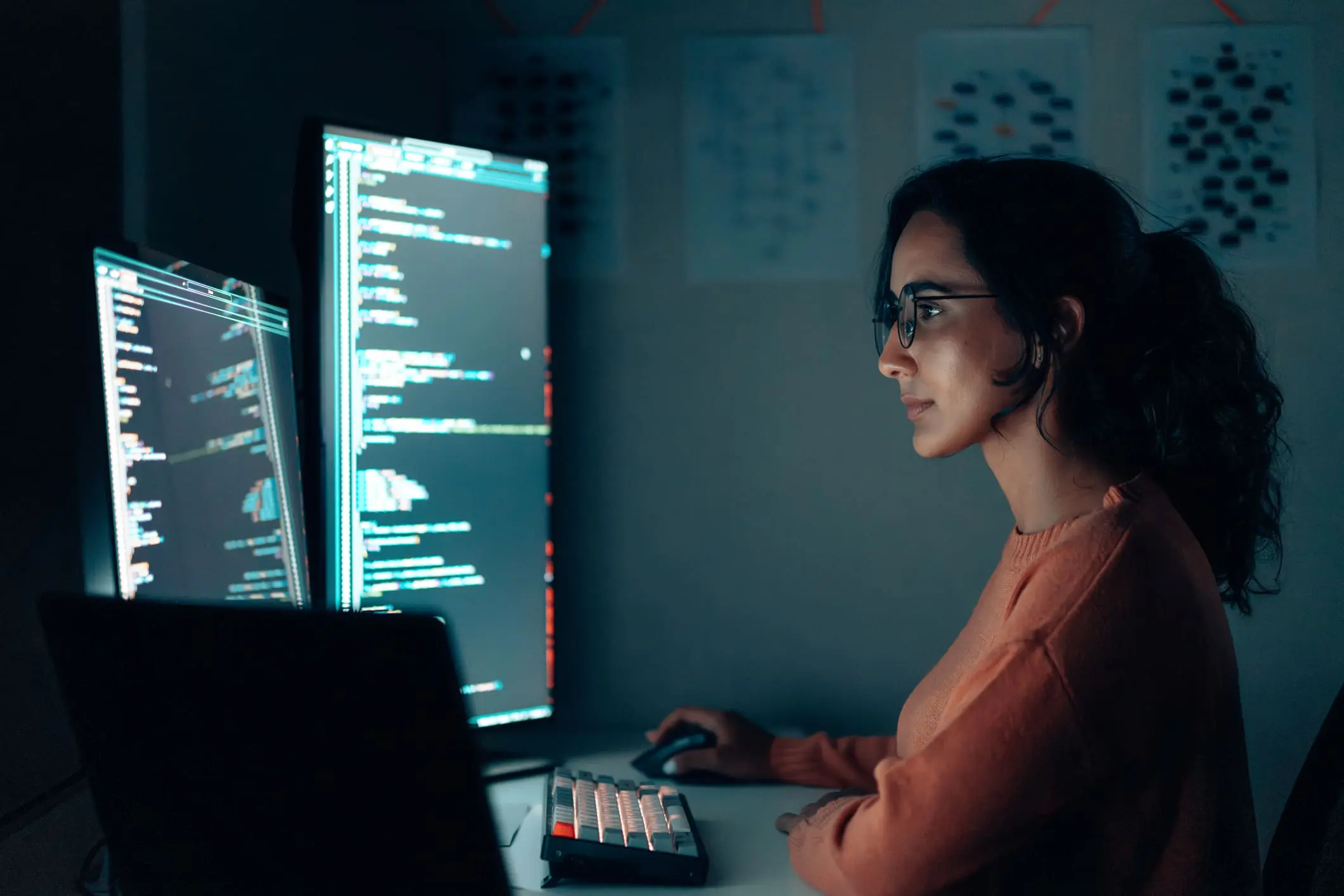 A woman with glasses and a ponytail, wearing an orange sweater, works on multiple computer screens displaying code in a dark room.
