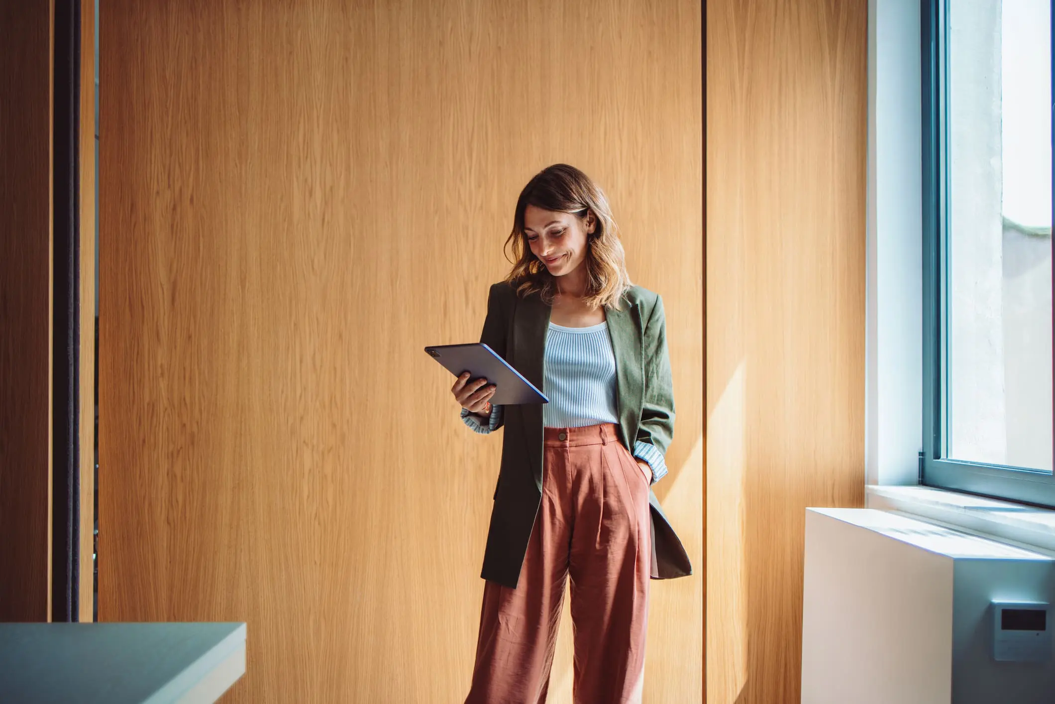 A woman in a green blazer and rust-colored pants stands in front of a wooden wall, smiling as she looks at a tablet.