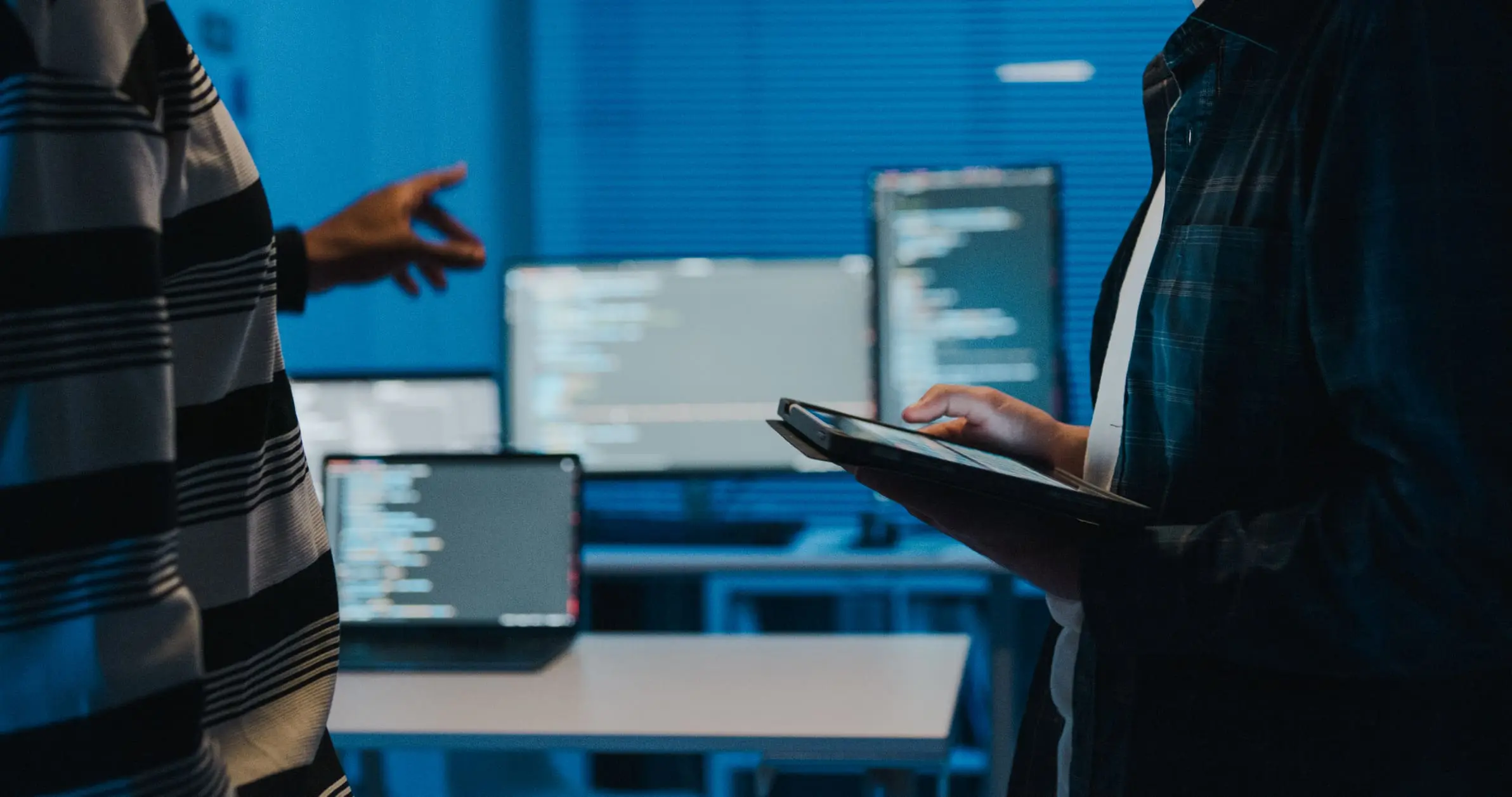 Two people in a blue-lit room, one pointing at a screen while the other uses a tablet. Computer monitors with code are visible in the background, suggesting a tech or programming environment.