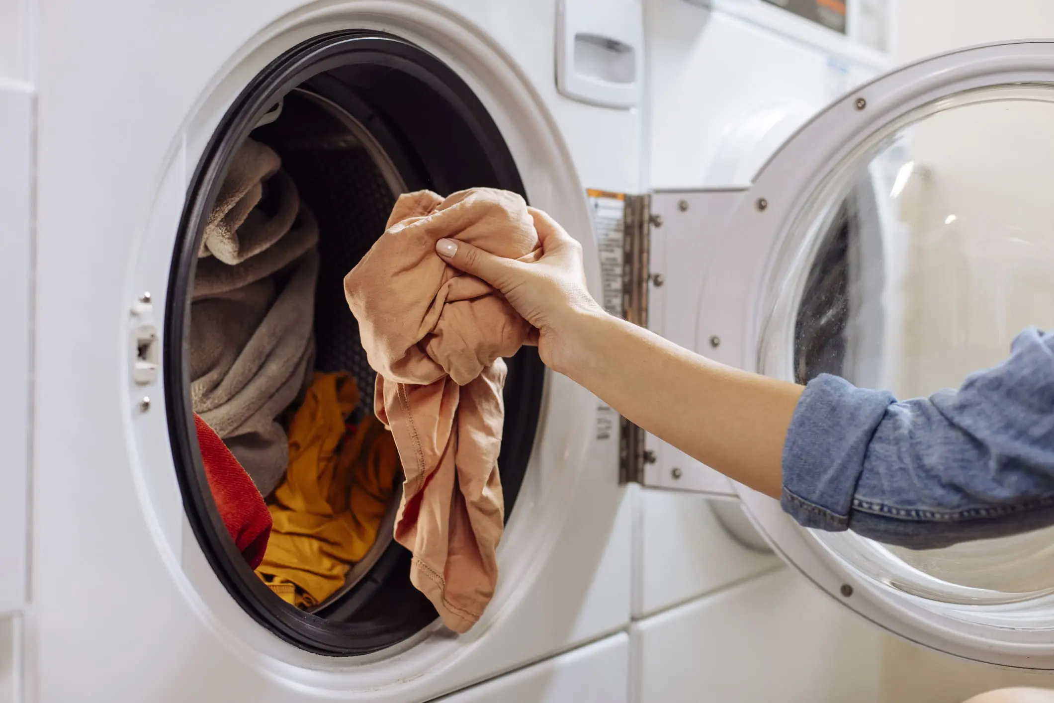 A hand puts a peach-colored garment into an open front-loading washing machine filled with other clothes.