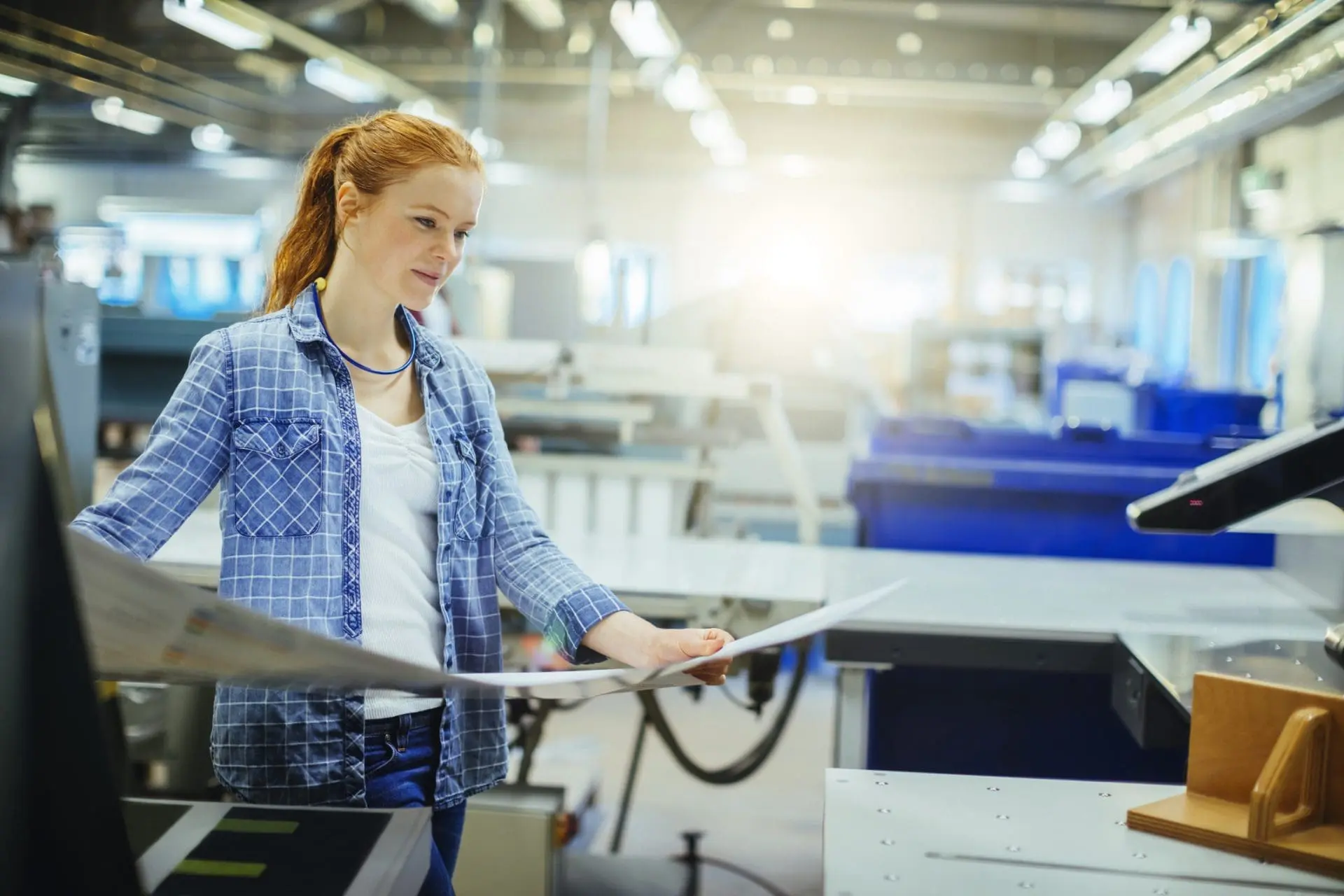 A young woman with red hair, wearing a plaid shirt, intently examines papers in a brightly lit industrial workshop.