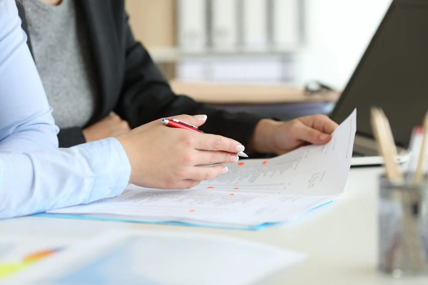 Close-up of two people's hands reviewing a document marked with red edits, one holding a red pen and the other holding the paper.