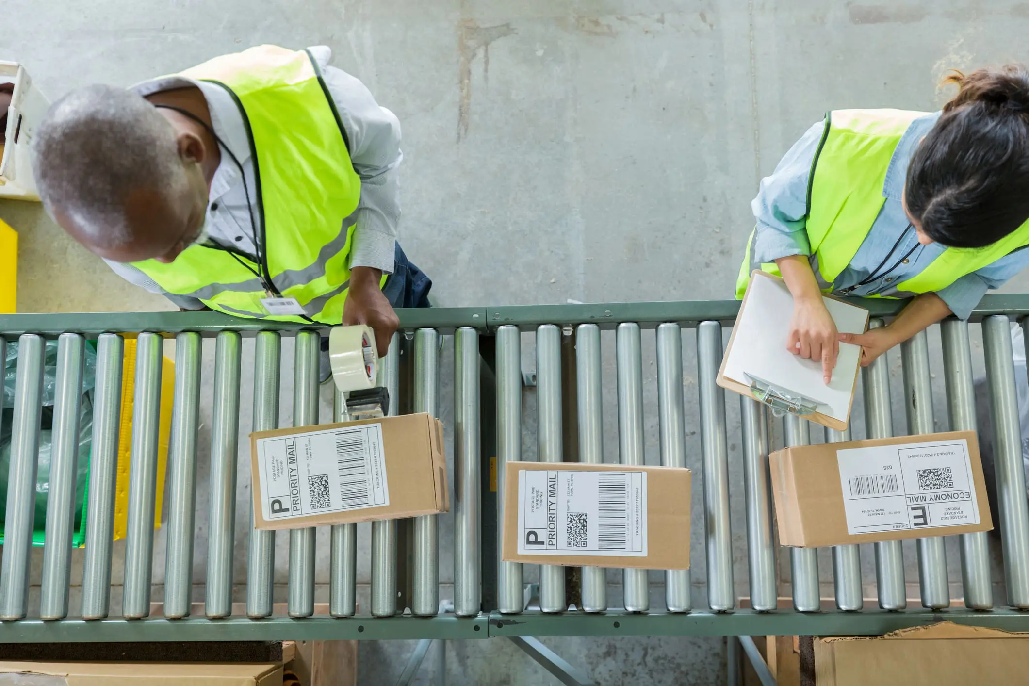 Overhead view of two workers in high-visibility vests processing packages on a conveyor belt in a warehouse. A man holds tape, and a woman holds a clipboard, with "PRIORITY MAIL" and "ECONOMY MAIL" boxes visible.