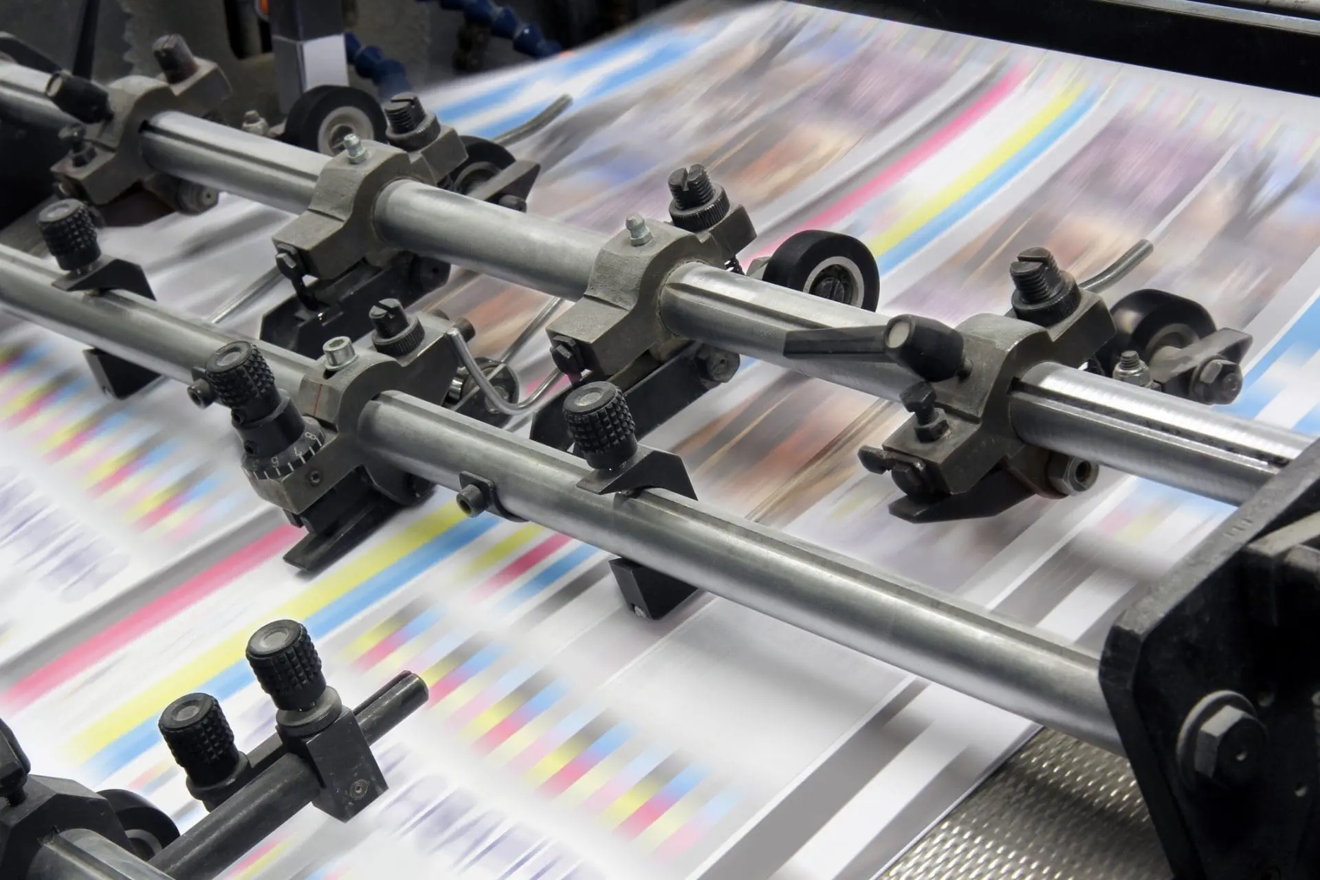 Close-up of an industrial printing press with paper moving rapidly through metal rollers. The paper shows blurred streaks of cyan, magenta, yellow, and black colors, indicating active printing.