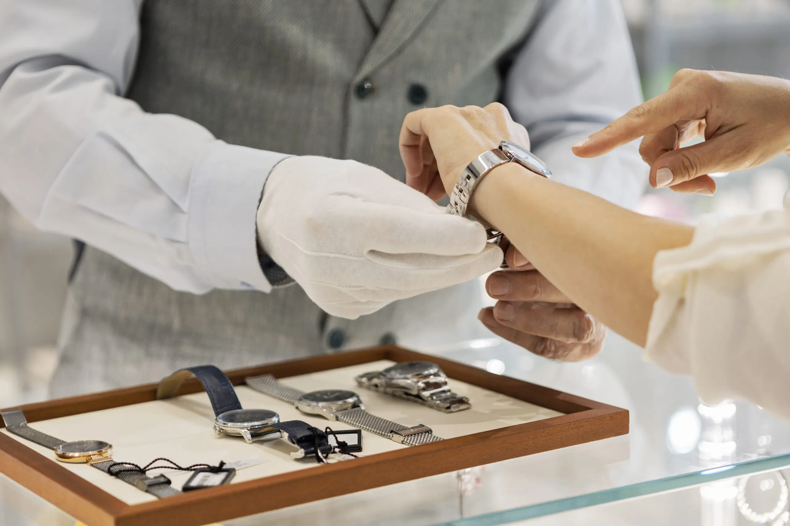 Adherent (formerly Compliance & Risks). A sales associate wearing white gloves helps a customer try on a silver watch in a store, with other watches displayed on a tray.