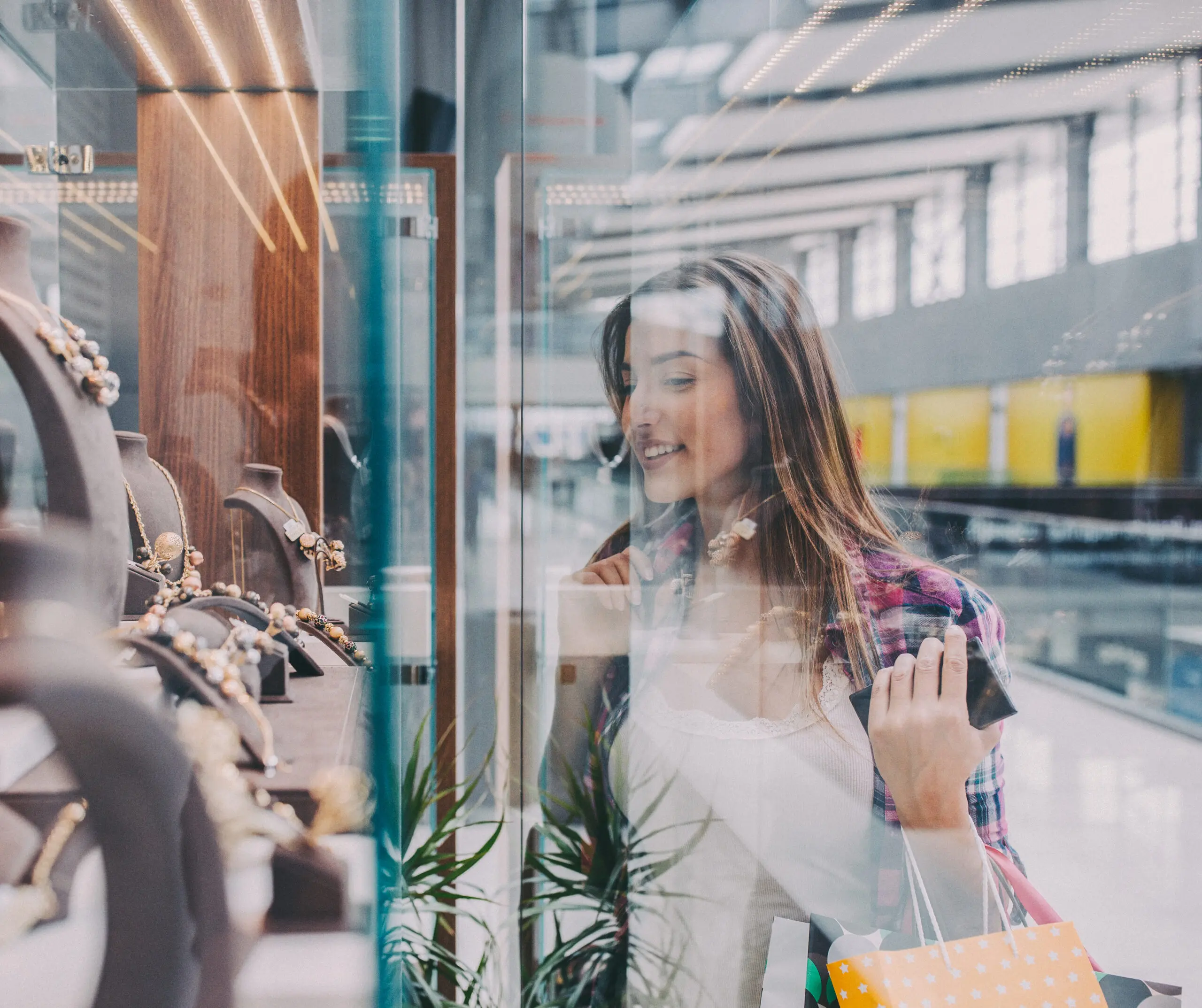 Adherent (formerly Compliance & Risks). A smiling young woman with long brown hair looks intently at a jewelry display in a store window, holding shopping bags.