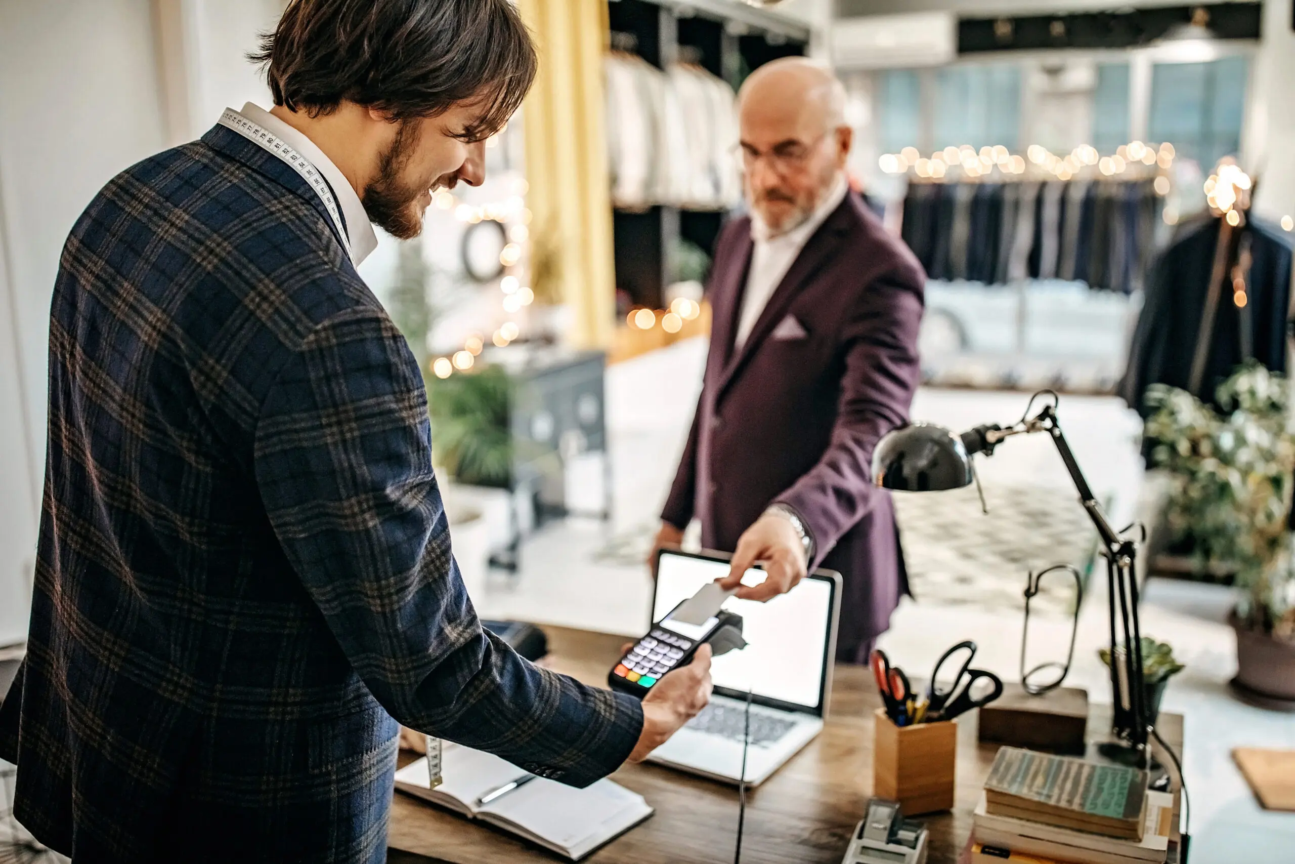 Adherent (formerly Compliance & Risks). A younger tailor in a plaid jacket holds a payment terminal while an older customer in a purple suit taps a card to pay. They are in a well-lit shop with clothes racks and a desk.
