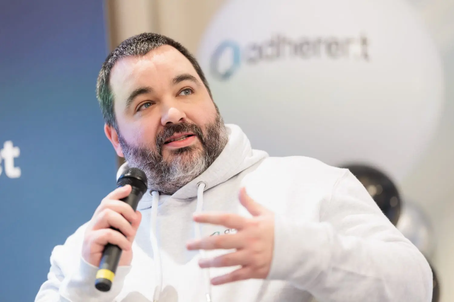 A man with a salt-and-pepper beard and light blue eyes speaks into a microphone, gesturing with his right hand. He wears a grey hoodie, and a blurred 'adherent' logo is in the background.