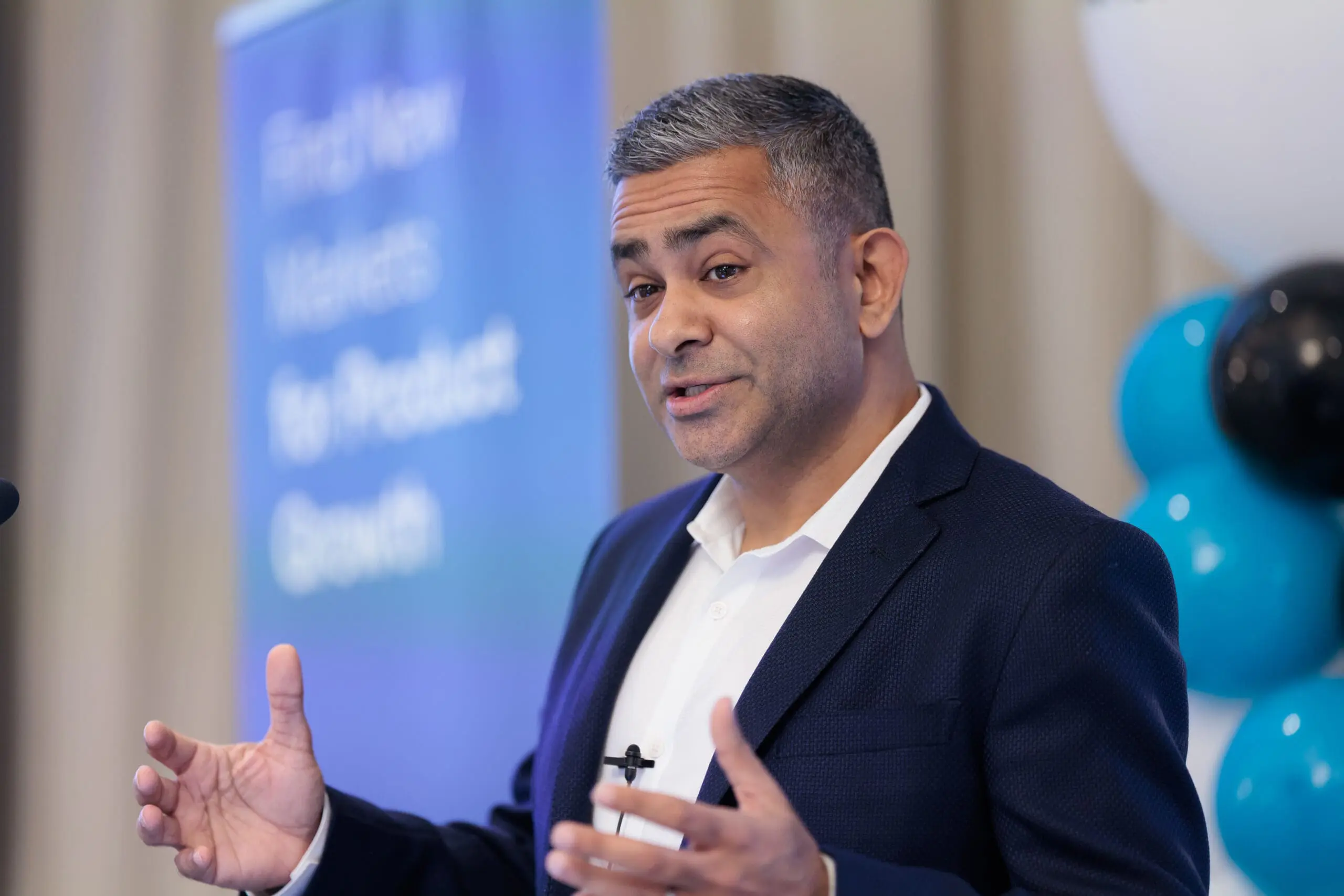 A man with grey-streaked dark hair, wearing a blue suit jacket and white shirt, speaks and gestures with his hand. A blurred blue banner with text 'Find New Markets For Product Growth' is behind him, along with blurred black and blue balloons.