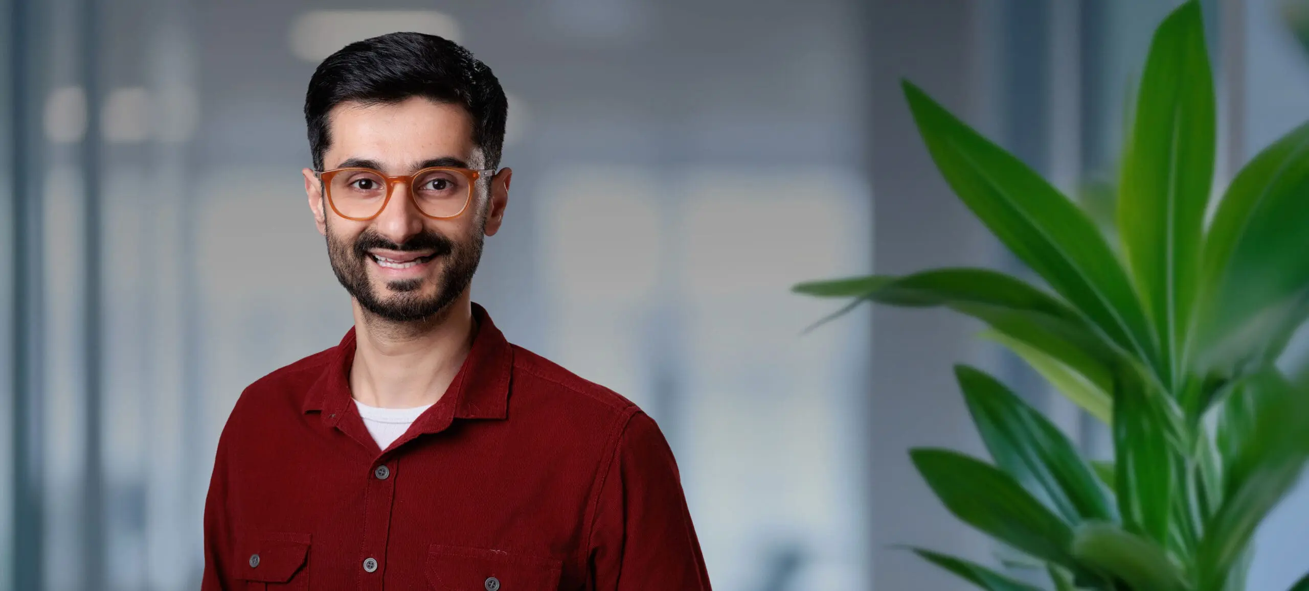 Adherent (formerly Compliance & Risks). A smiling man with dark hair, a beard, and amber glasses wears a red corduroy shirt in a blurred office setting with a green plant.