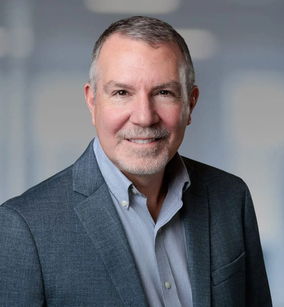 Headshot of a smiling man with grey hair and beard, wearing a grey blazer and blue shirt, in a blurred office background with a green plant.