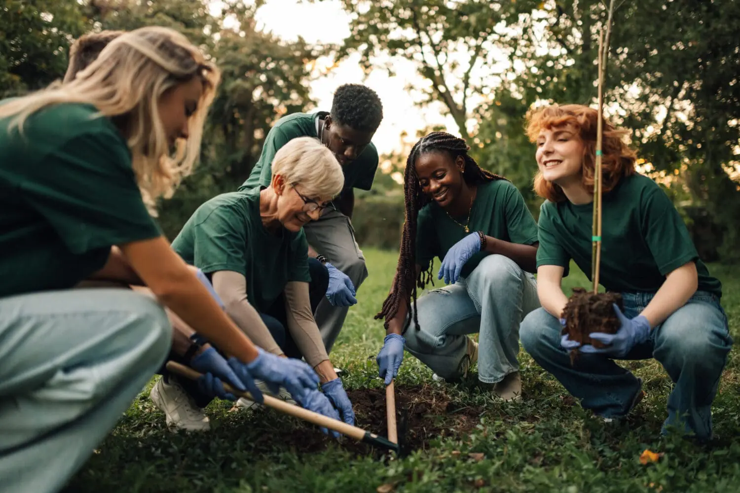 Five diverse volunteers in green shirts and blue gloves smiling and working together to plant a young tree in a grassy outdoor setting.