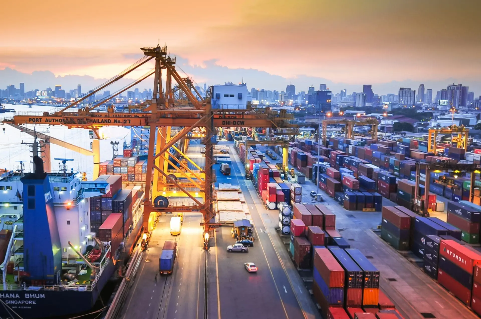 Aerial view of a busy port at dusk, with a container ship, large yellow gantry cranes, rows of stacked shipping containers, and a city skyline in the background under an orange and purple sky.