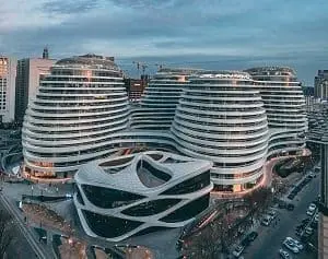 Aerial view of a complex of modern, white, curvilinear buildings with horizontal bands and a smaller, angular building in the foreground, set in a city landscape at dusk.