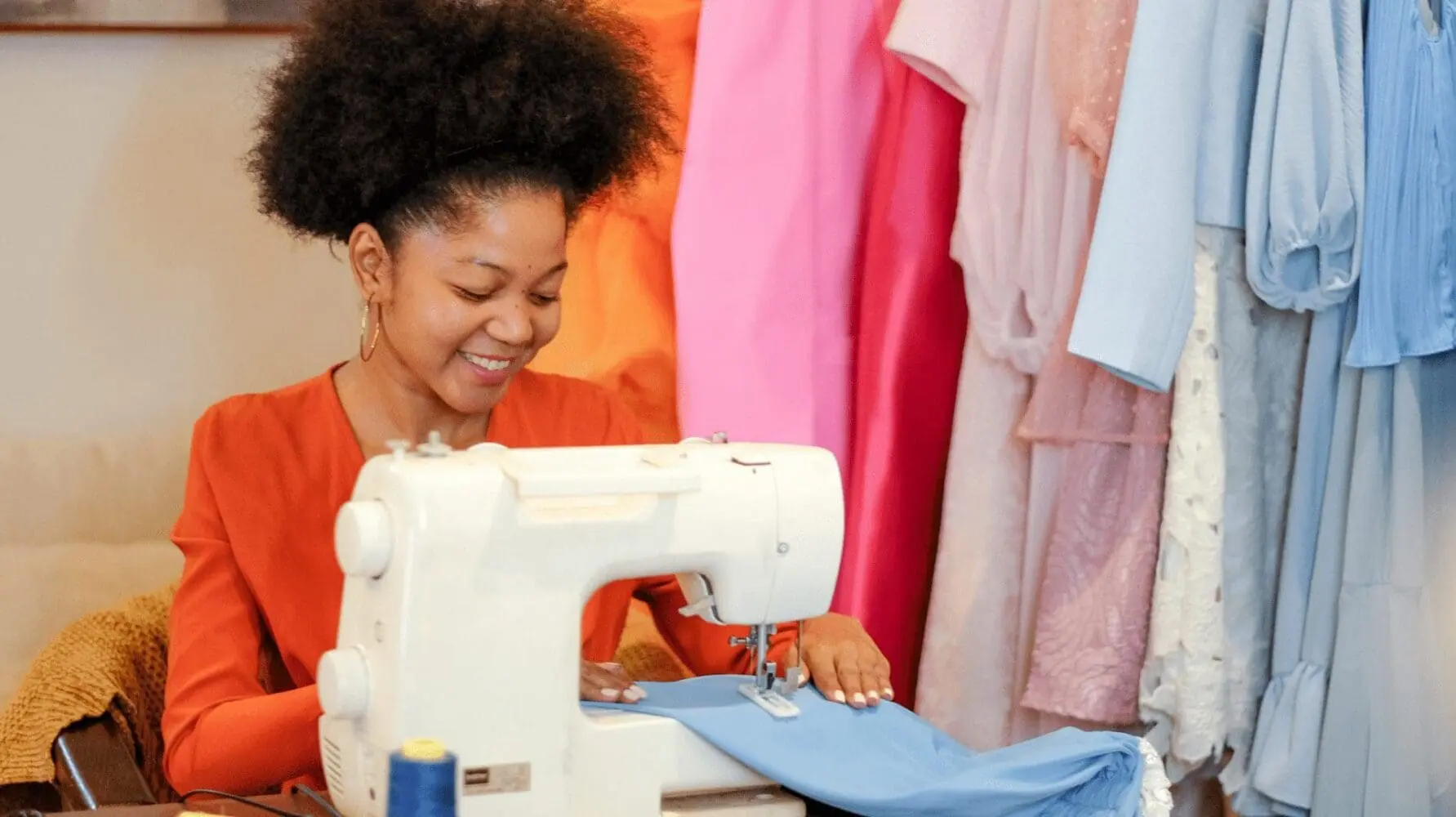Adherent (formerly Compliance & Risks). A smiling woman with dark, curly hair sewing light blue fabric on a white sewing machine, with colorful clothes hanging in the background.