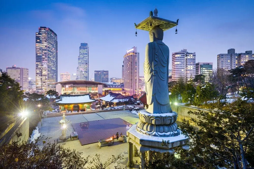 Adherent (formerly Compliance & Risks). A large stone Buddha statue seen from behind, overlooking a traditional temple complex covered in snow, with a modern city skyline illuminated at dusk.