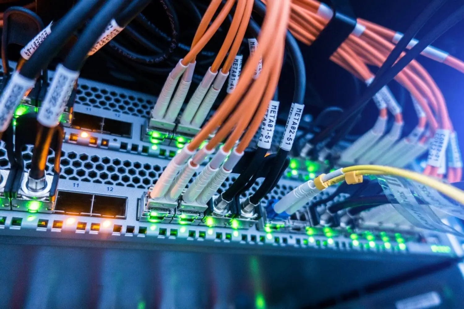 Adherent (formerly Compliance & Risks). Close-up of network equipment with numerous orange fiber optic cables, black cables, and a yellow cable plugged into ports. Many green and some orange indicator lights are glowing on the silver hardware. Labels like 'META01-49' are visible on the cables.