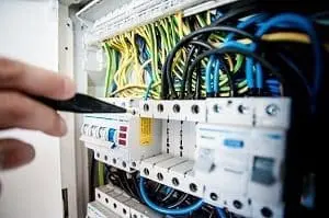 Adherent (formerly Compliance & Risks). Close-up of a hand holding a black tool near an open electrical panel with white circuit breakers and colorful wires.