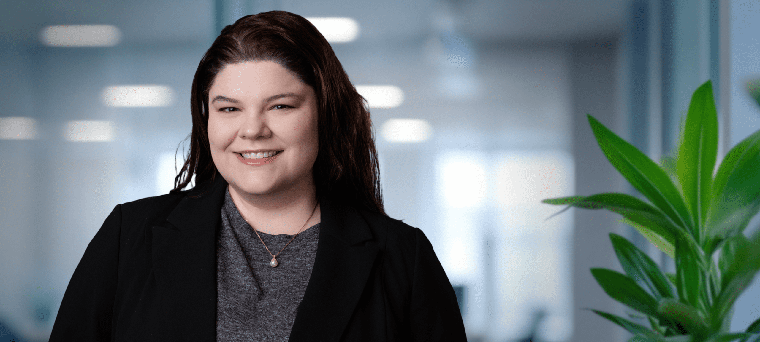 Adherent (formerly Compliance & Risks). Headshot of a smiling woman with dark hair, wearing a black blazer and a pearl necklace, against a blurred office background.