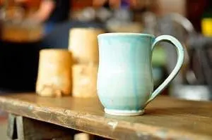 Adherent (formerly Compliance & Risks). Light blue ceramic mug with a handle on a dark wooden table, with blurred beige objects in the background.