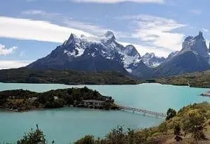 Adherent (formerly Compliance & Risks). A panoramic view of a turquoise lake with an island connected by a bridge, set against a backdrop of towering, snow-capped mountains under a partly cloudy sky.