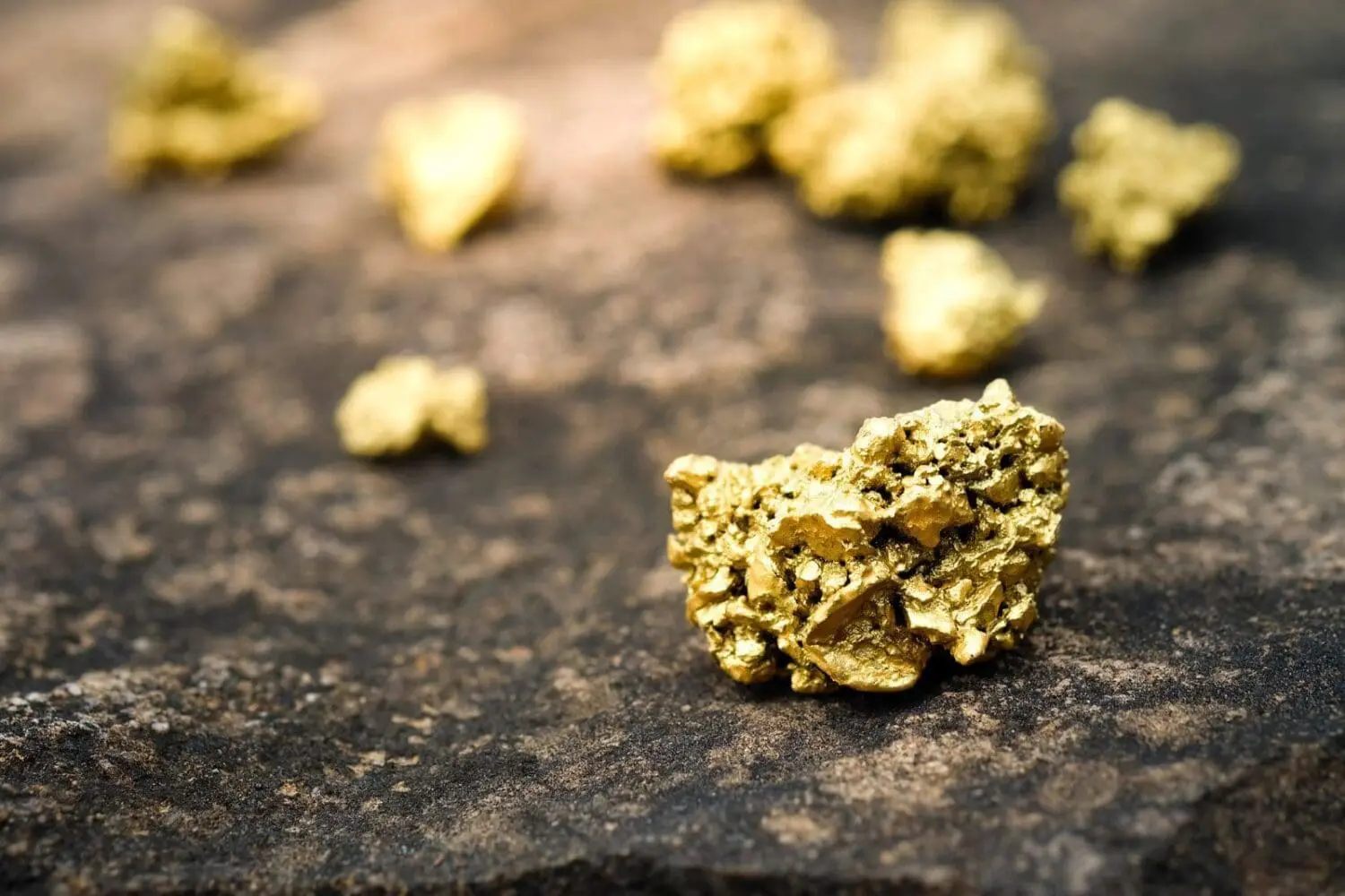 Adherent (formerly Compliance & Risks). Several raw gold nuggets of various sizes scattered on a dark, textured rock surface. The foreground nugget is in sharp focus, showing its rough, bright yellow, metallic texture.