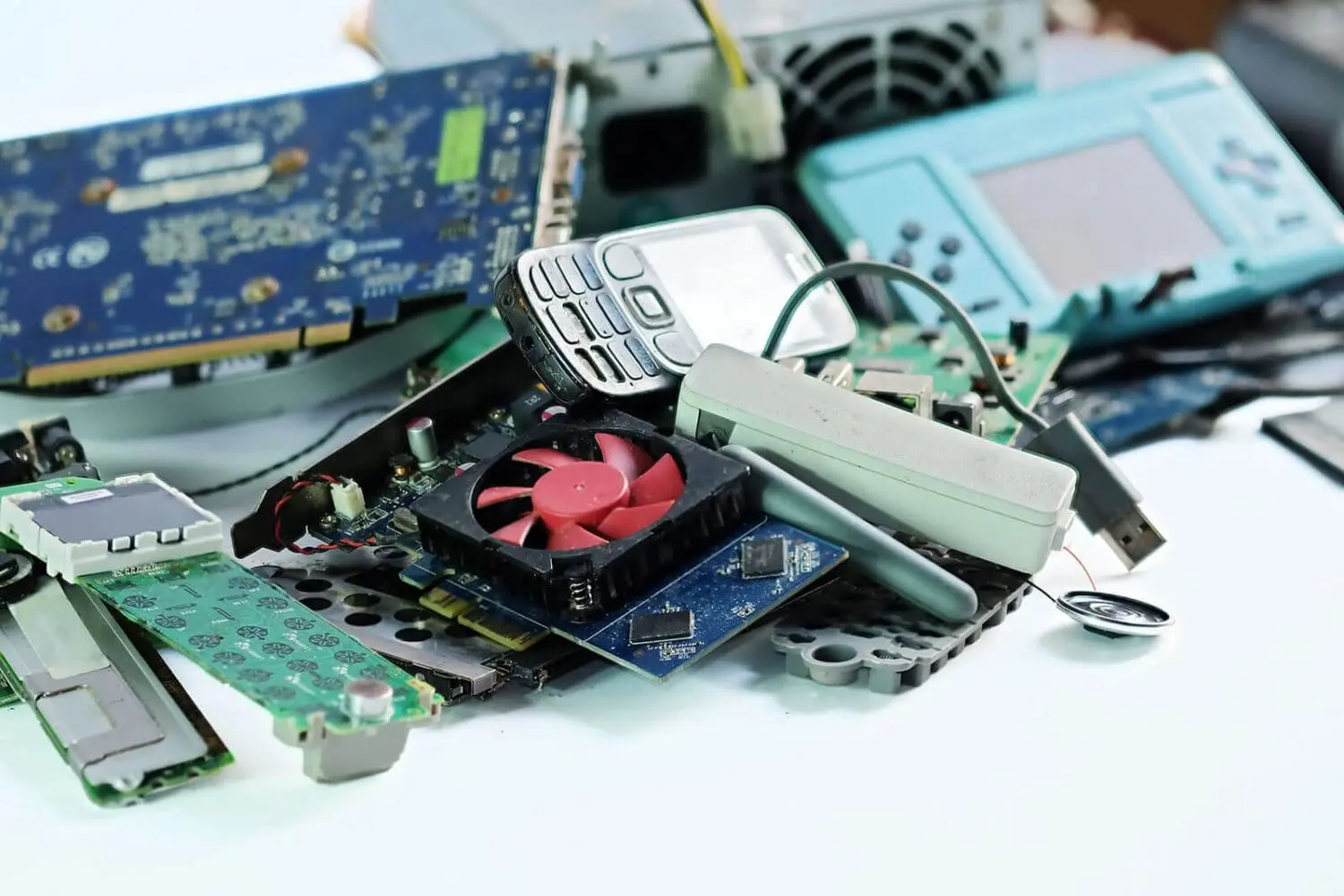 Adherent (formerly Compliance & Risks). A pile of electronic waste including circuit boards, an old mobile phone, a red cooling fan, and a handheld gaming device.