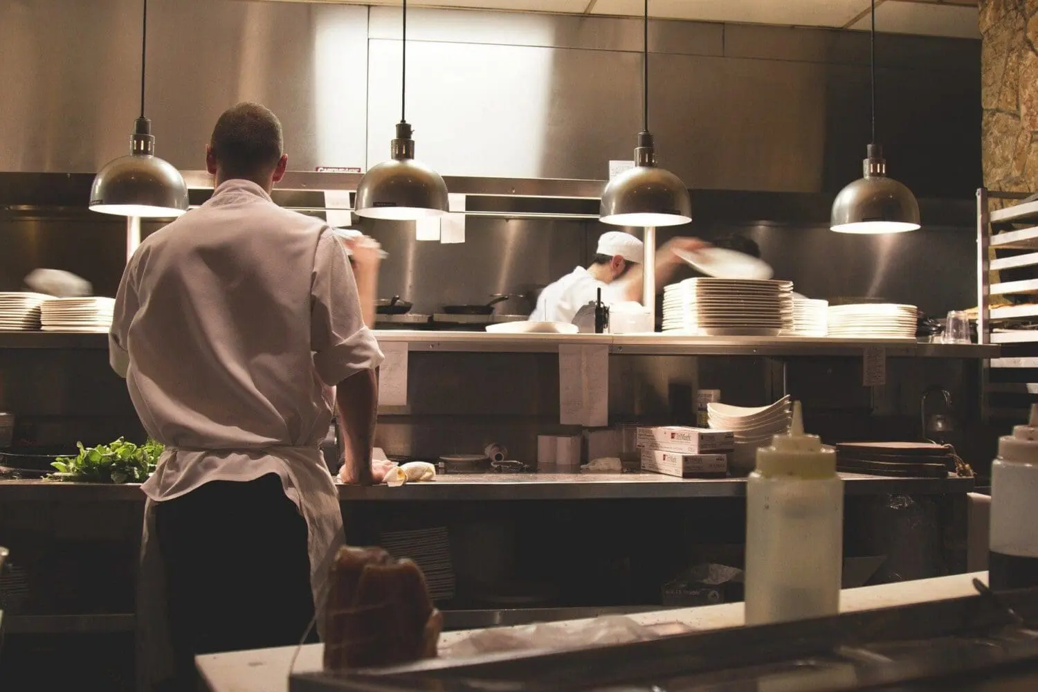 Adherent (formerly Compliance & Risks). A chef in a white jacket stands with their back to the camera in a professional kitchen, with another chef and stacked plates in the background under warm lighting.